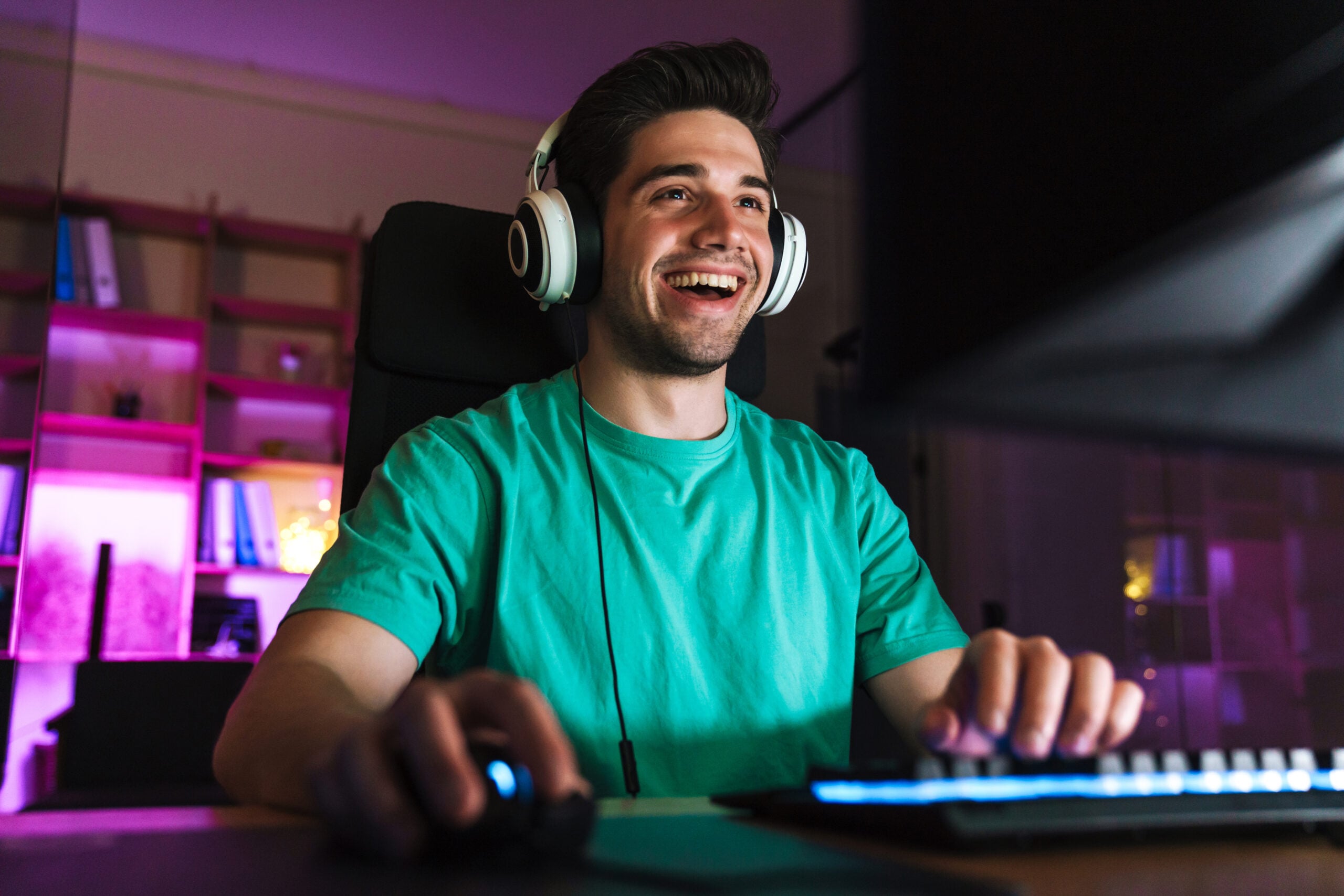 Image of caucasian happy man in headphones playing video game on computer while sitting at table indoors
