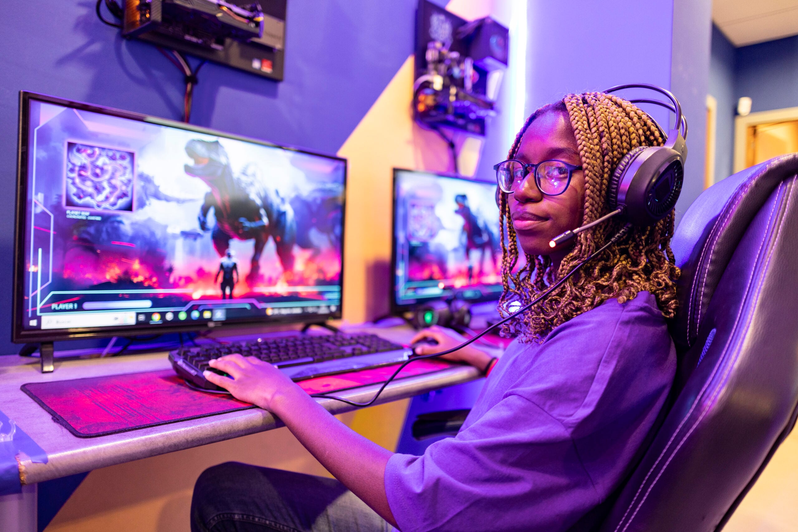 Young woman wearing headset and playing a futuristic online video game fighting a dinosaur using her computer and mouse in a purple gaming room