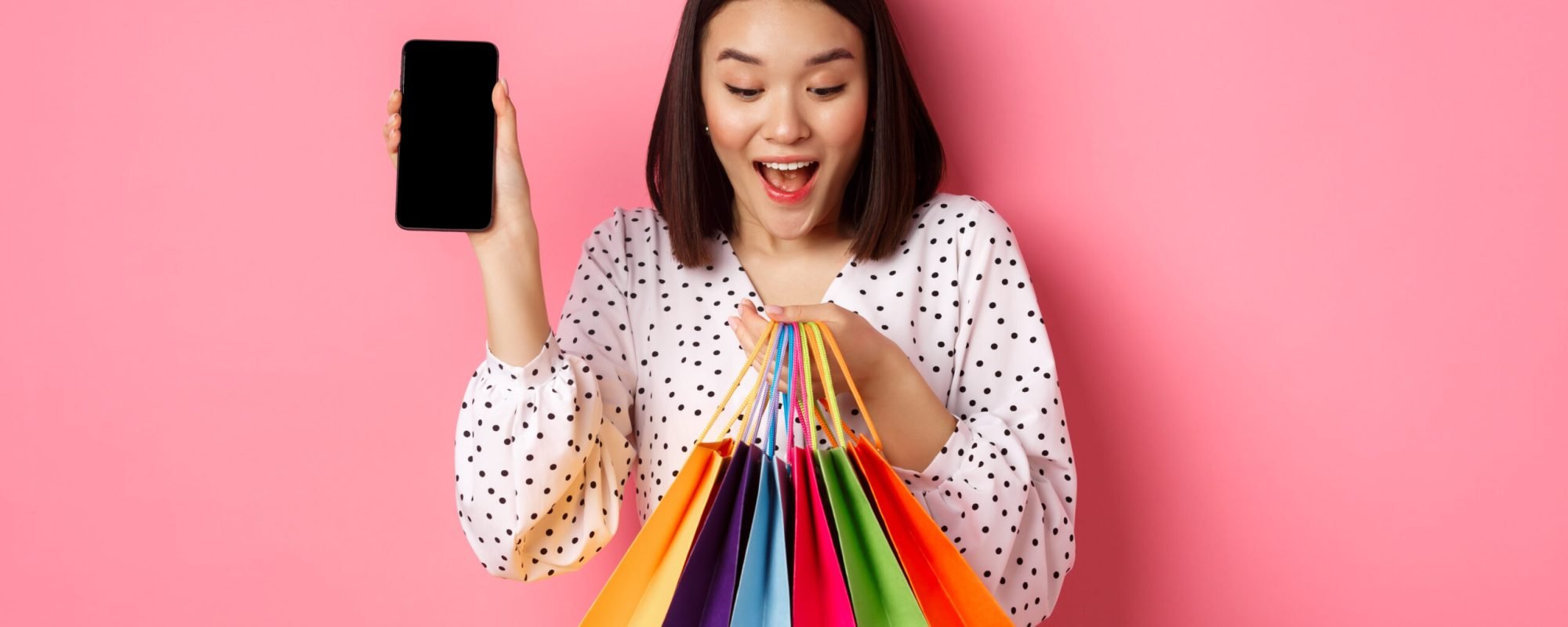 Attractive asian woman showing smartphone app and shopping bags, buying online via application, standing over pink background.