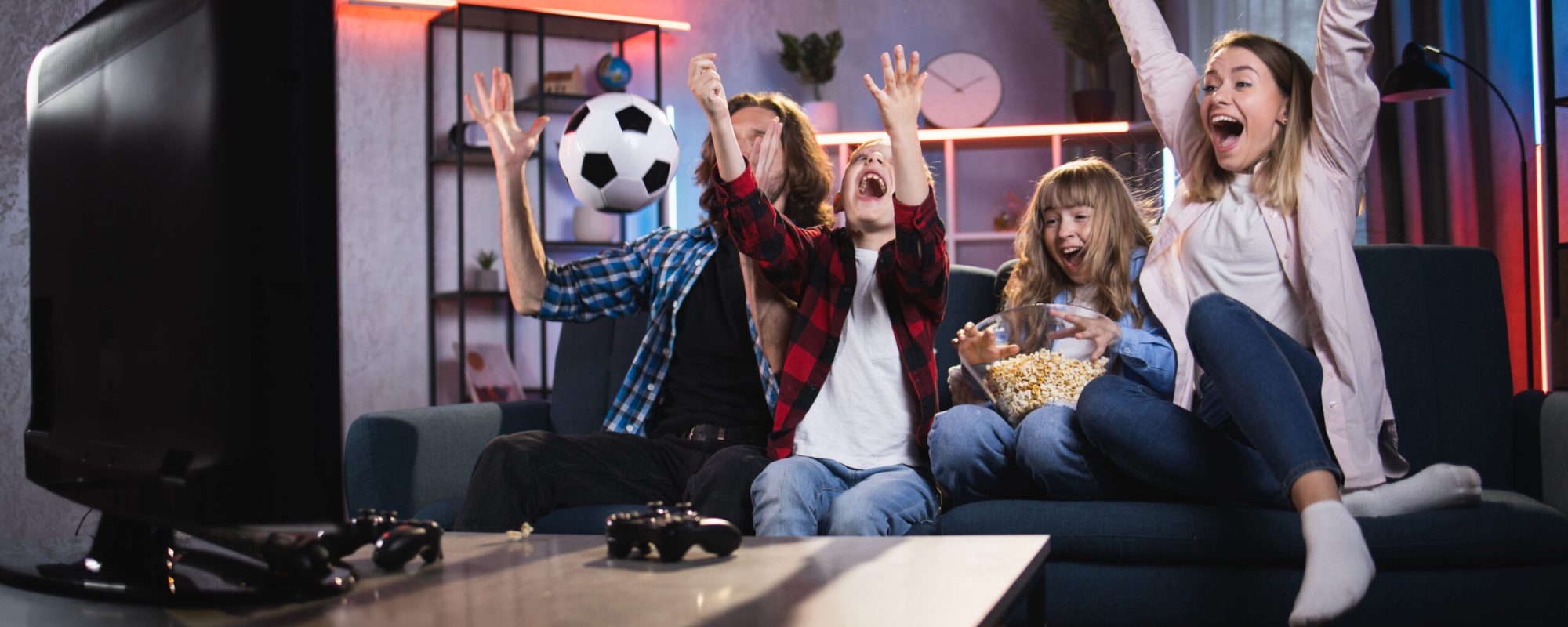 Emotional parents with little son and daughter celebrating goal of favorite team during soccer match. Satisfied family sitting on couch and watching sport competition on TV.