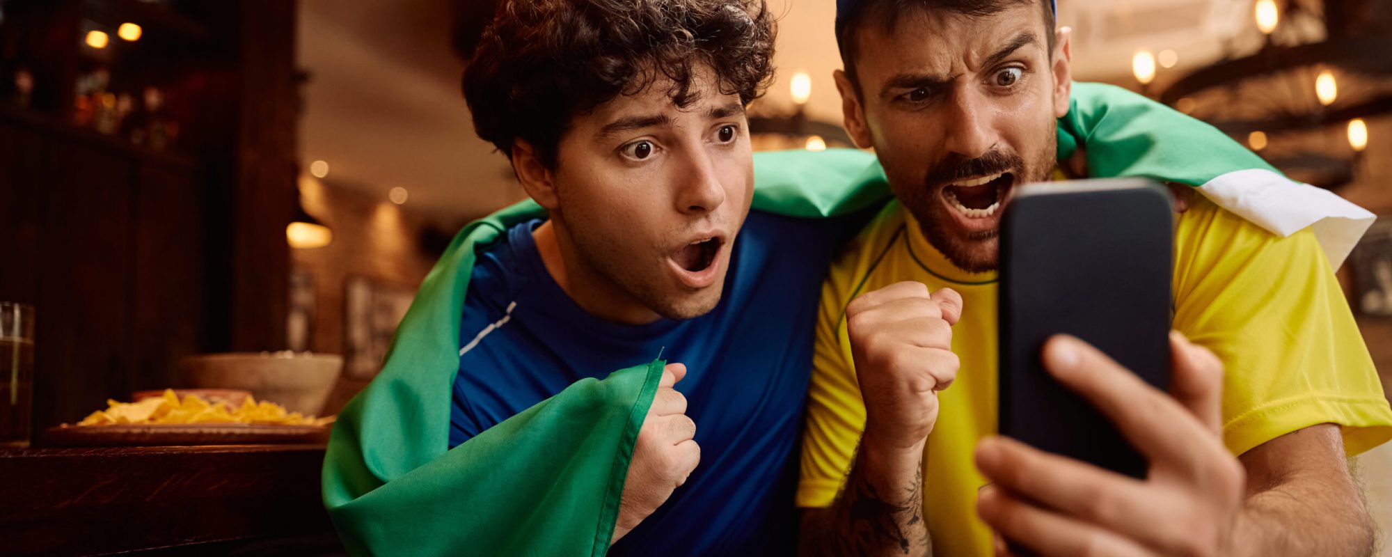 Young men cheering while watching sports match on smart phone in a pub.