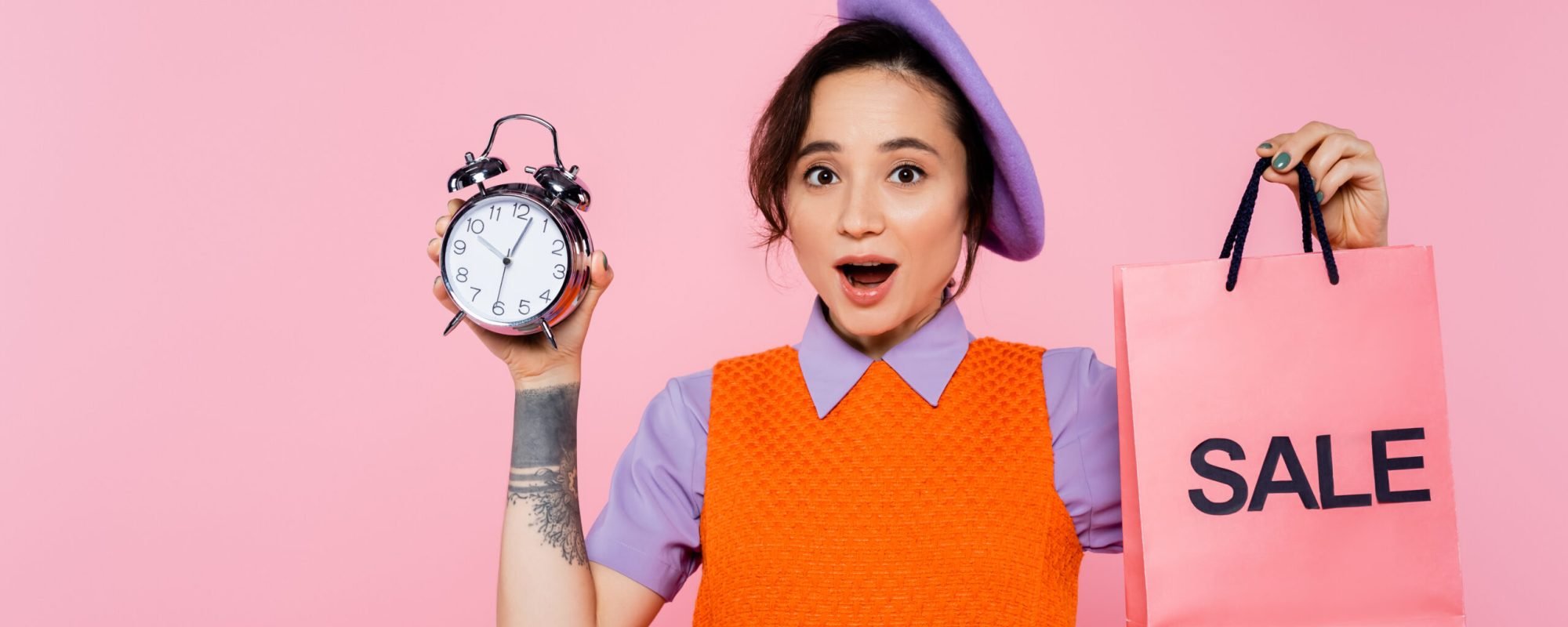 excited woman in bright clothes holding vintage alarm clock and sale shopping bag isolated on pink
