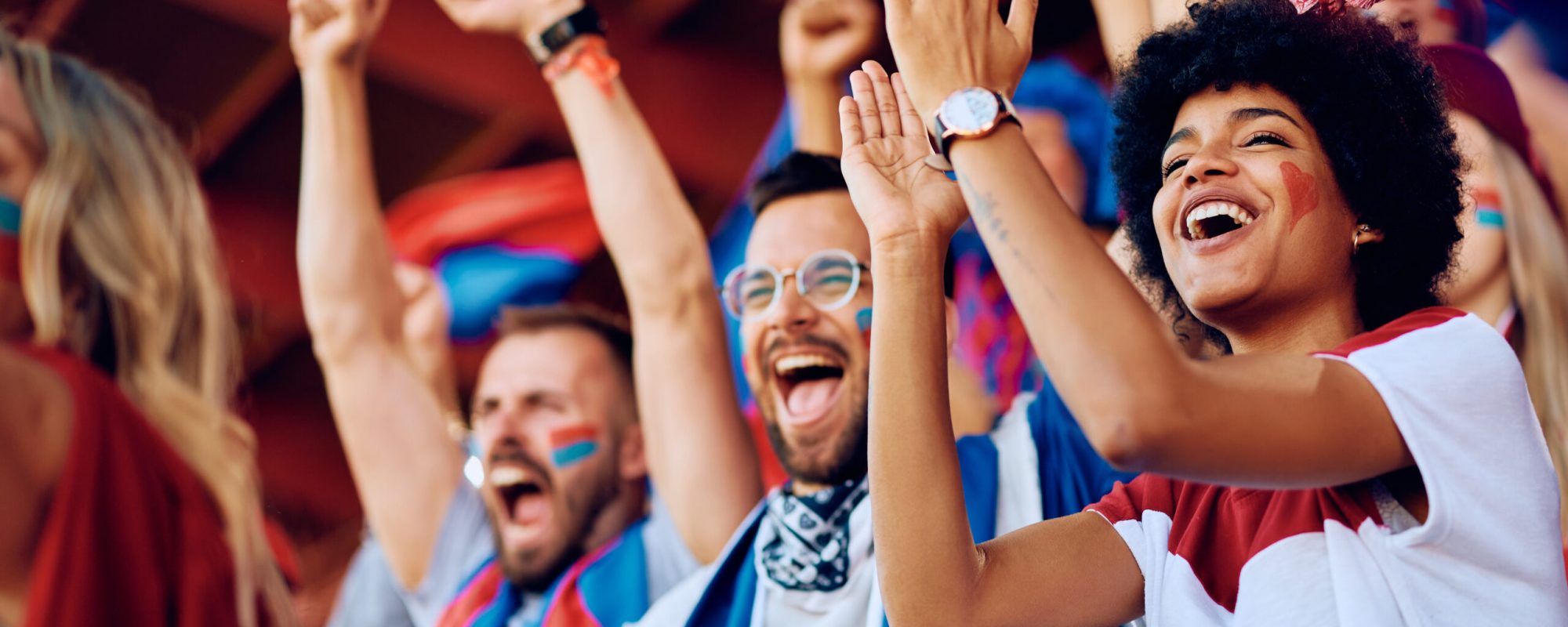 Group of cheerful sports fans celebrating victory of their favorite team while spectating game from stadium stands. Focus is on African American woman.