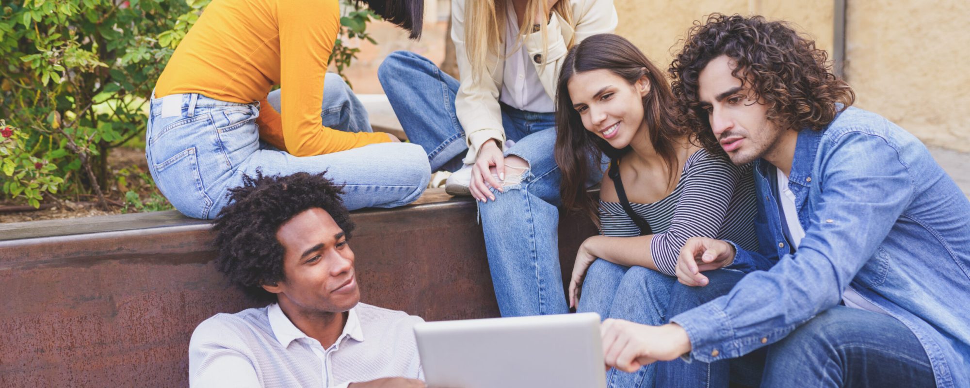 Multi-ethnic group of students looking at something on a digital tablet sitting outdoors.