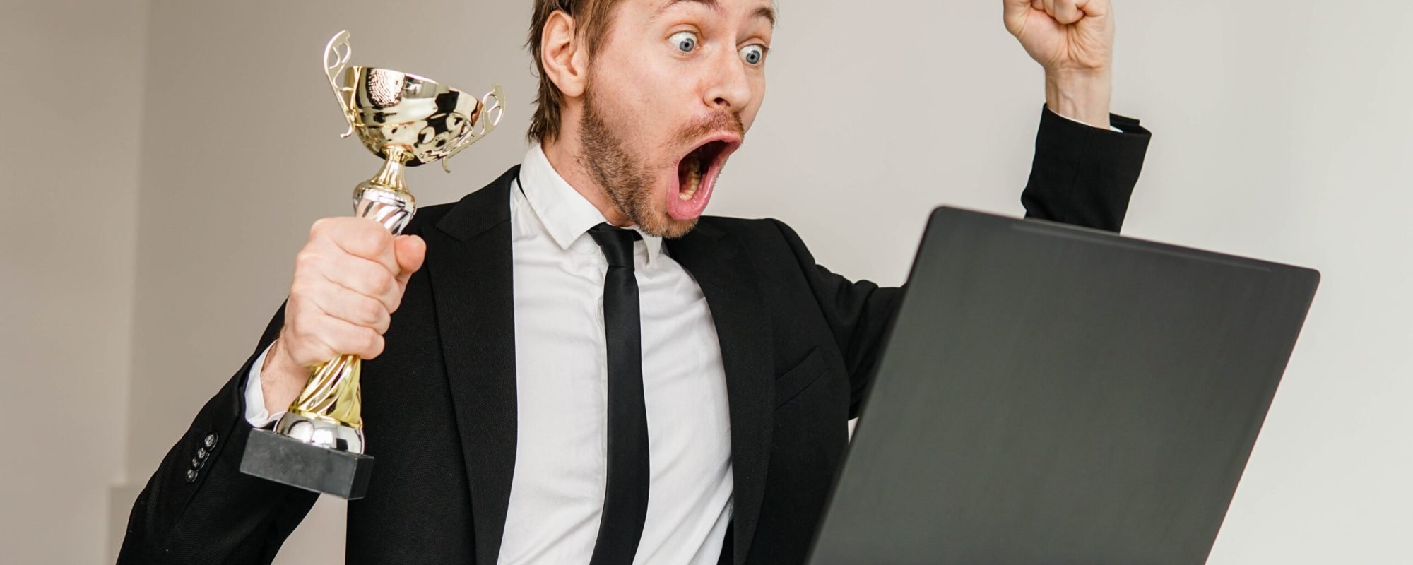 Successful businessman in suit celebrating victory with golden cup trophy while watching online ceremony on laptop.